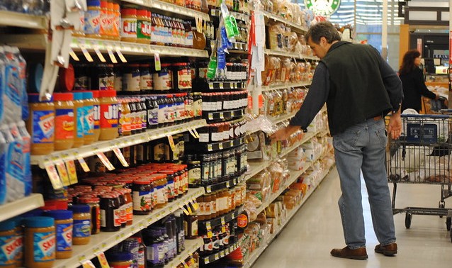 Man in grocery aisle with peanut butter and jam, looking at bread 
