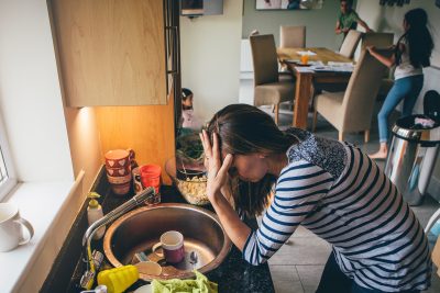 Stressed mum at home. She has her head in her hands at a messy kitchen sink and her children are running round in the background.