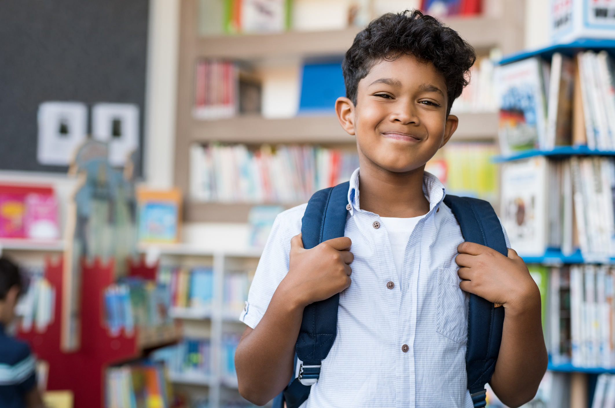 Portrait of smiling Latino boy looking at camera, carrying backpack and standing in library at school.