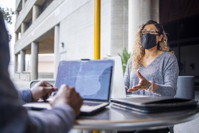 Woman sitting at desck across from man with Laptops in front of them. Woman is facing camera wearing a face mask.