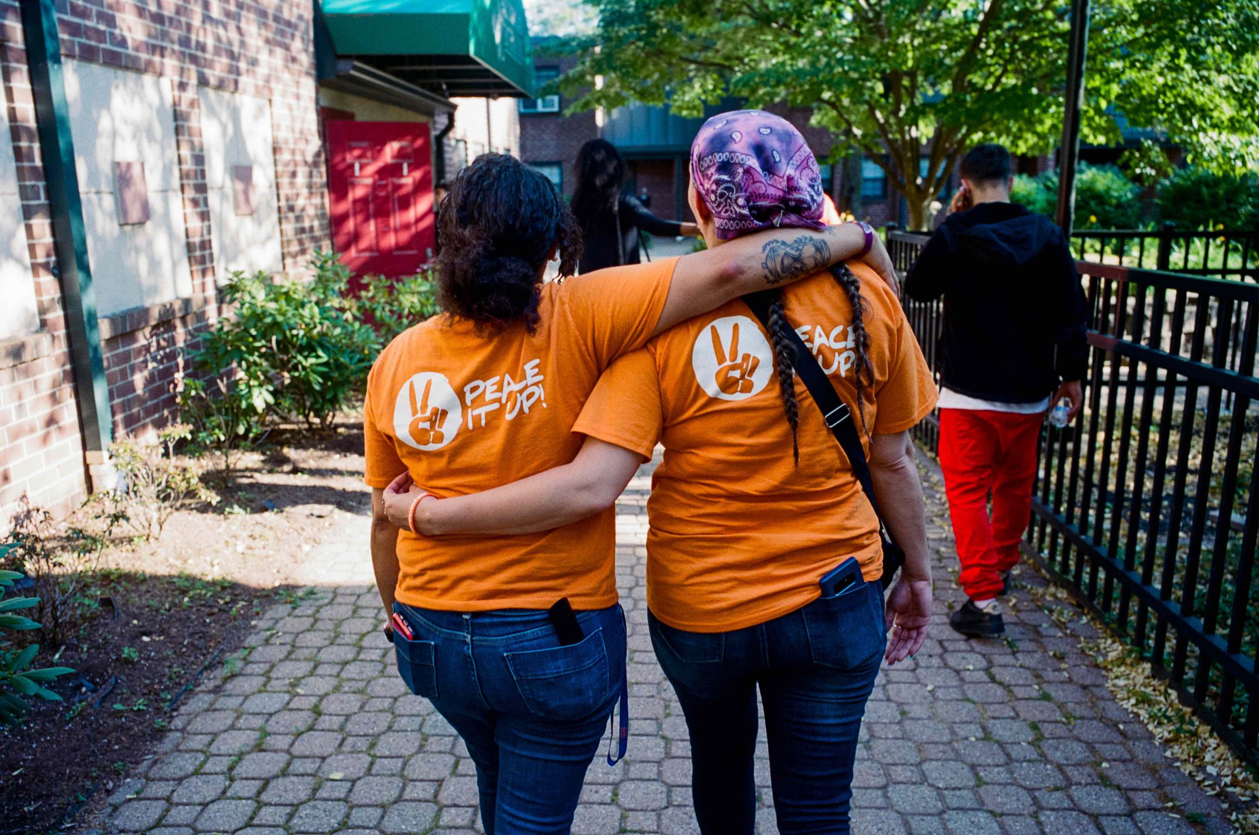 two women walking away from camera wearing jeans and orange t-shirts with Peace it up written on the back
