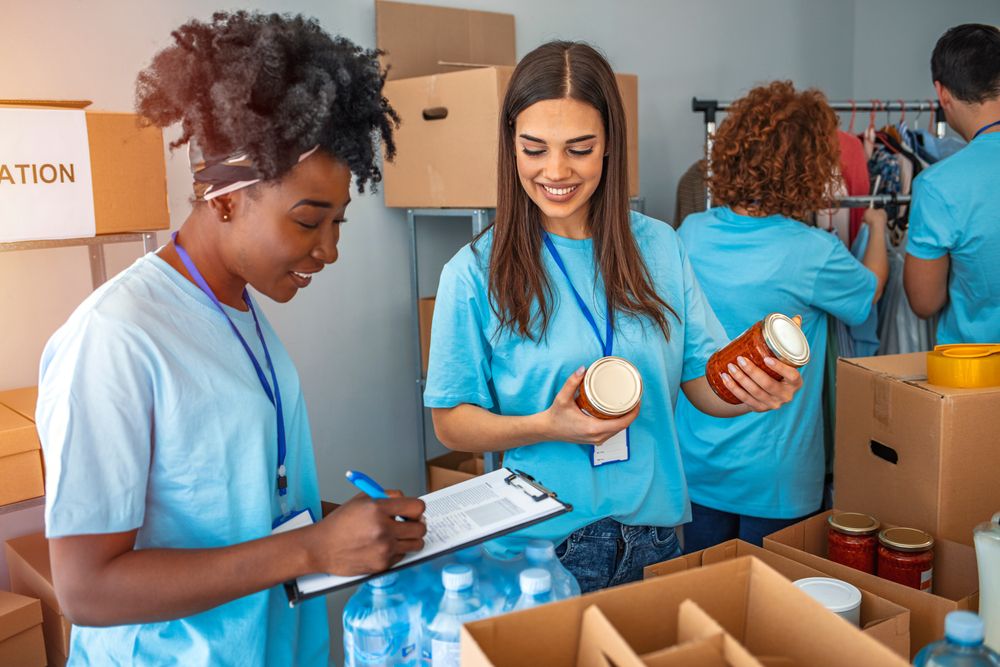 Two women, one Black and one White, volunteering in a food pantry pack boxes for distribution to hungry people. The Black woman holds a clipboard and is smiling; the White eoman holds two jars of tomato sauce.