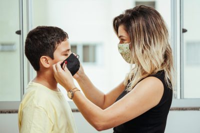 Mother wearing white and green cloth face mask faces son while putting on black coth fask mask on this face.