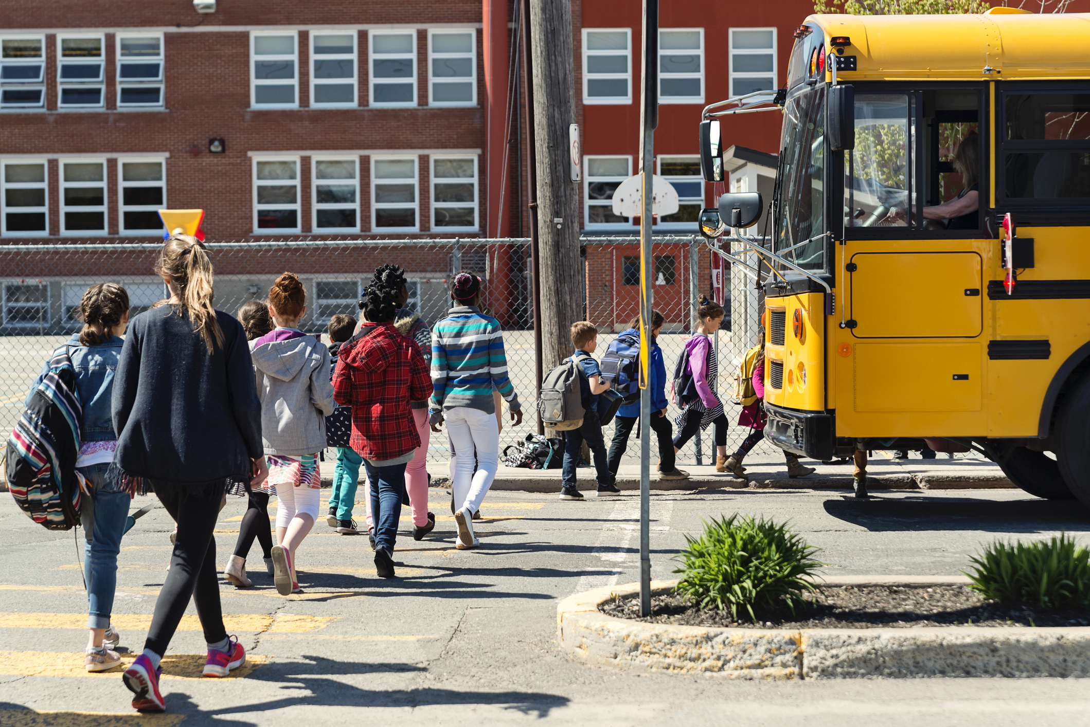 Kids in line crossing street to get on school bus in front of school building.