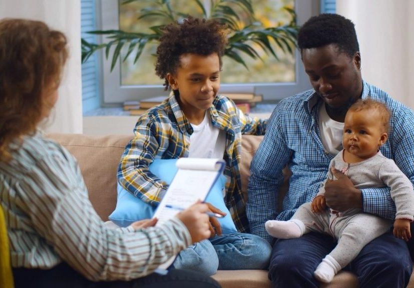White woman with notepad and pen sits with back to the camera, interviewing two Black adolescent boys with slight smiles wearing plaid shirts. One of them is holding a baby in his lap.