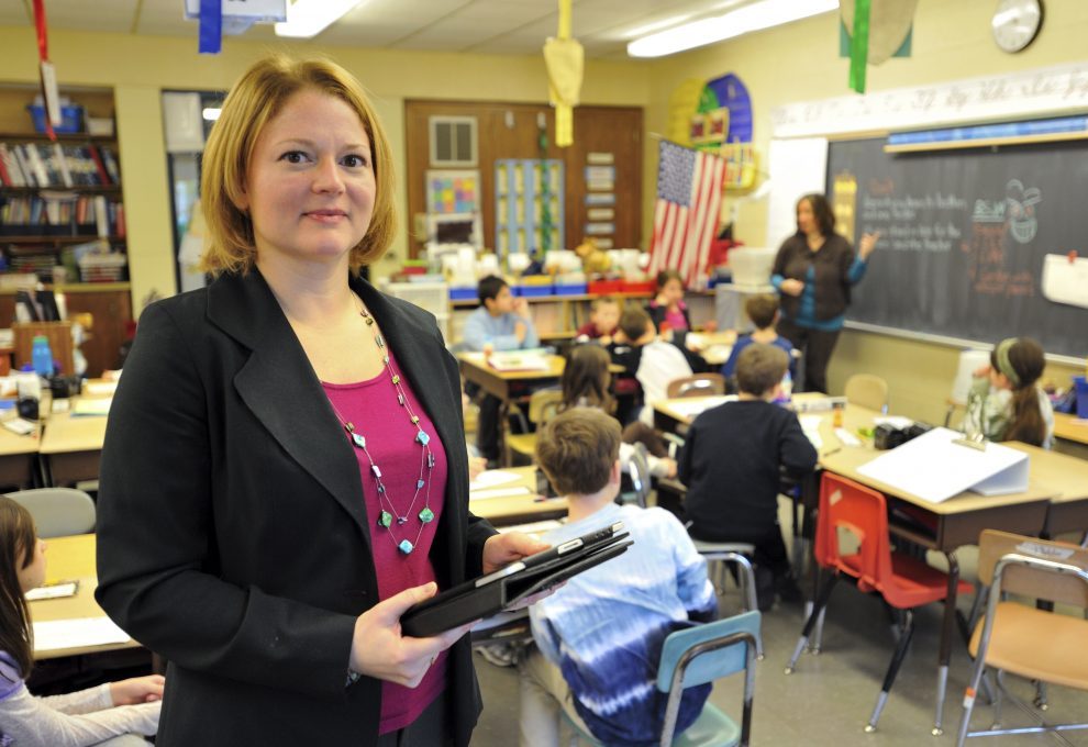 Sandra Chafouleas stands in front of a classroom of kids holding a binder and iPad. She is a white eoman with blonde hair wearing a raspberrry shirt with black blazer.