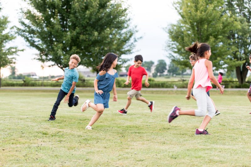A diverse group of elementary school students play tag outside at recess. They are running around the grass outside of the school.