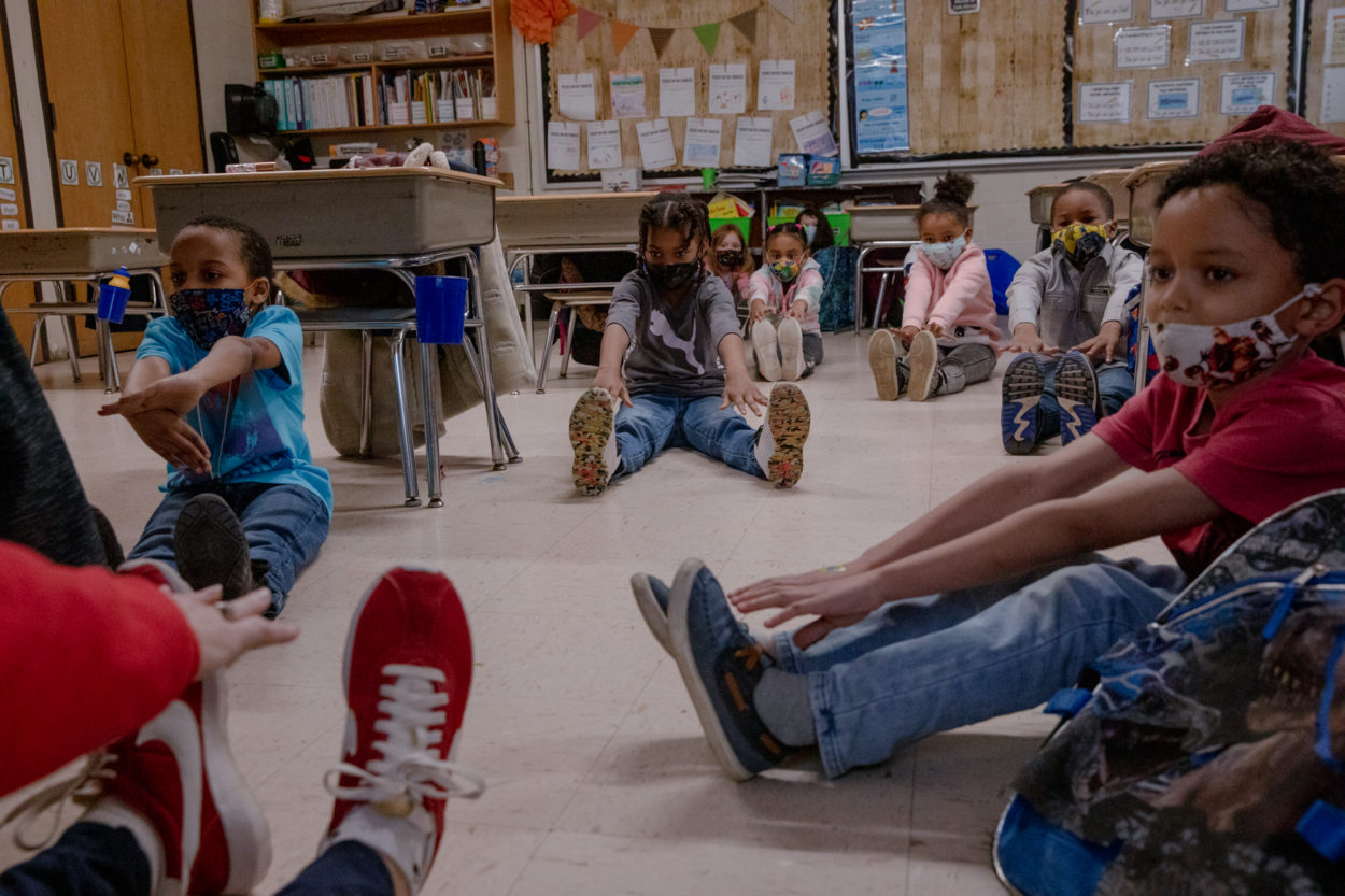 Group of young, diverse kids in school sit on the floor with legs stretched out and touching their toes. They are wearing masks