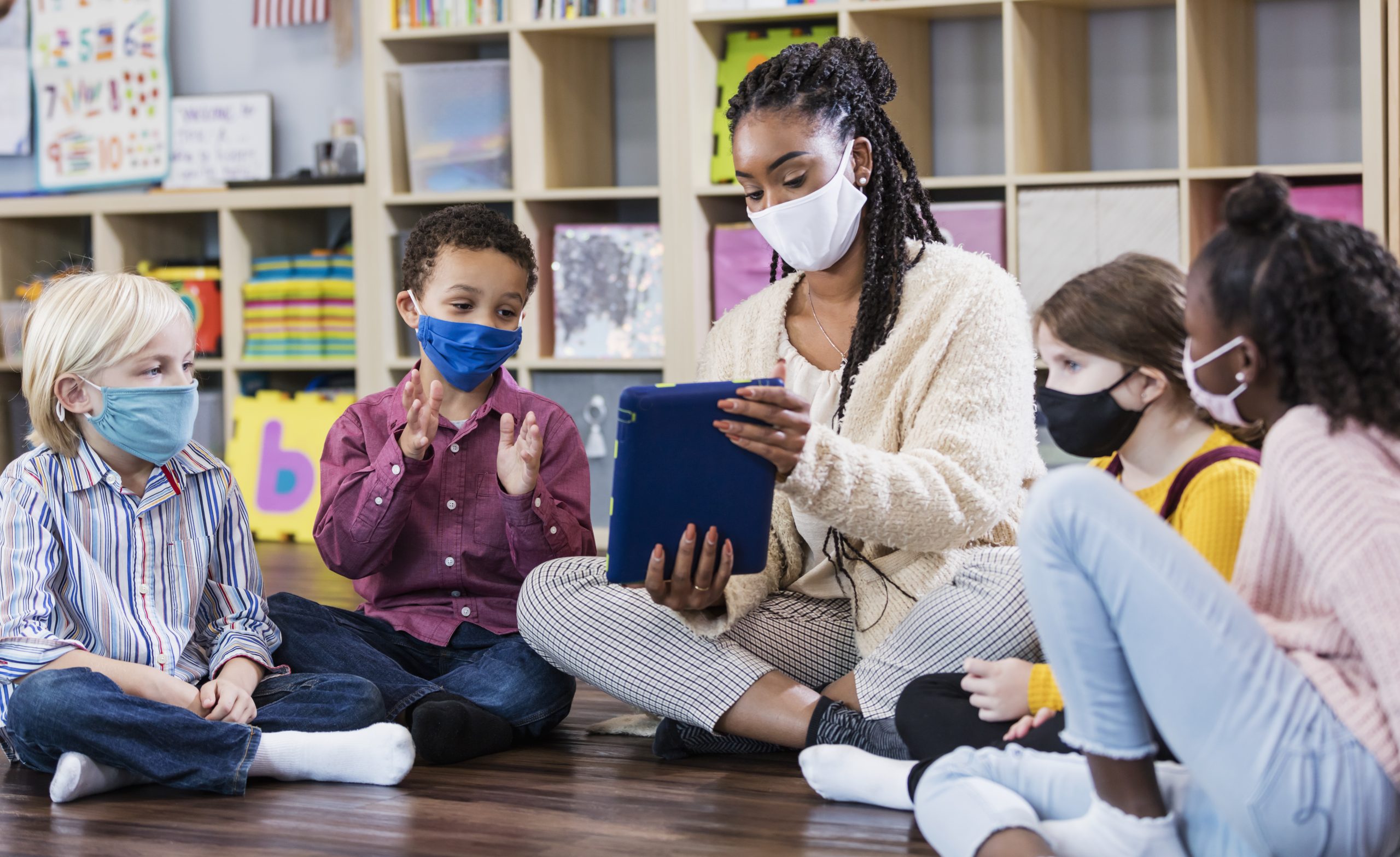Black woman teacher sits cross-legged with diverse children in school classroom. All of them are wearing masks. The teacher shows one student something on a tablet.