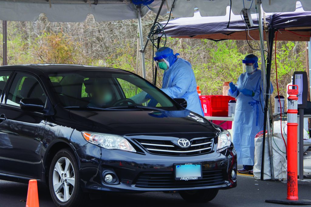 A black Toyota sedan pulls up to two health workers in PPE under a tent administering drive through COVID-testing