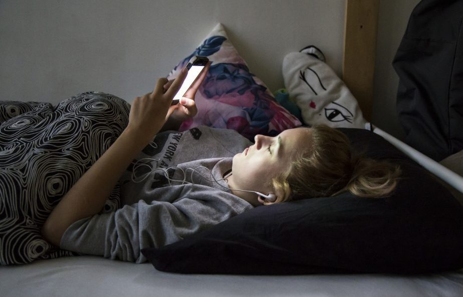 Teenager is lying on bed, looking mesmerized at her phone in dar