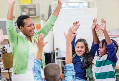 A Black female teacher sitting in a classroom with six elementary school students. They are sitting in chairs arranged in a circle, playing a game, stretching their arms in the air.