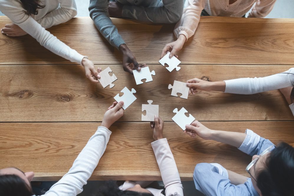 group of seven diverse adult hands holding white puzzle pieces toward each other