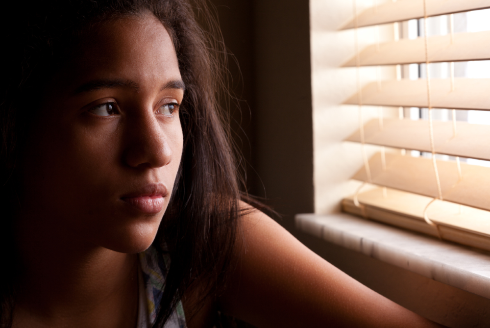 A Black teen girl stares out of a window through blinds with a somber expression on her face