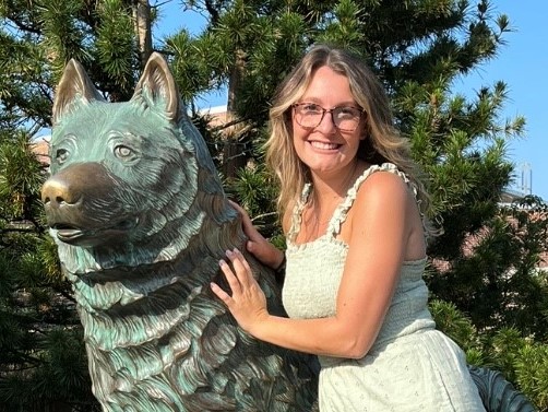Emma Zuk stands to the right of the UConn Husky statue. She is wearing glasses and in a white dress.