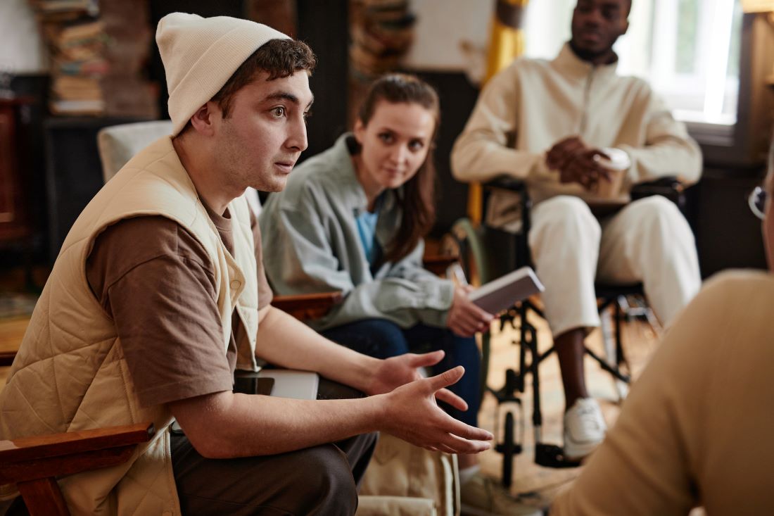 A teenager sits in a group of teens. He is wearing a white knit cap and down vest. He is gesturing with his hands.