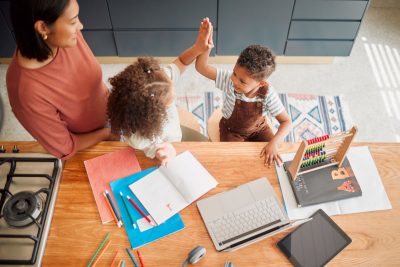 Young boy and girl sit at desk and give each other a high five while a teacher looks on.