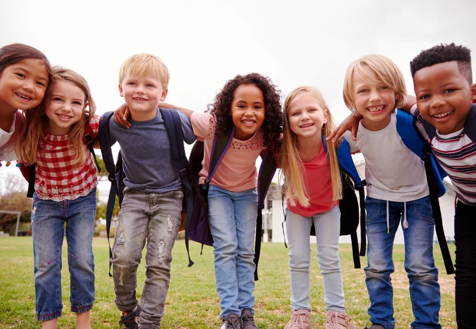group of smiling diverse kids on field of grass. They are wearing t shirts and jeans and have their arms around each other as they smile for the camera.