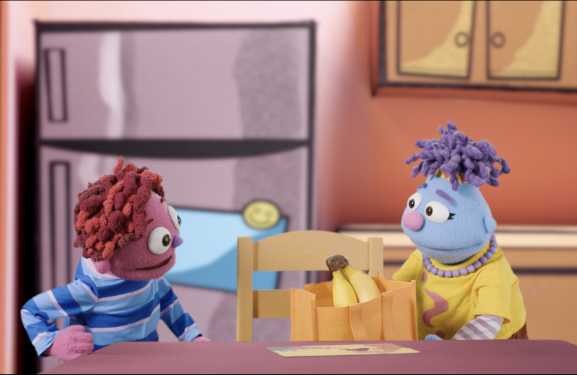 Two puppets stand at a kitchen table with a grocery bag of bananas