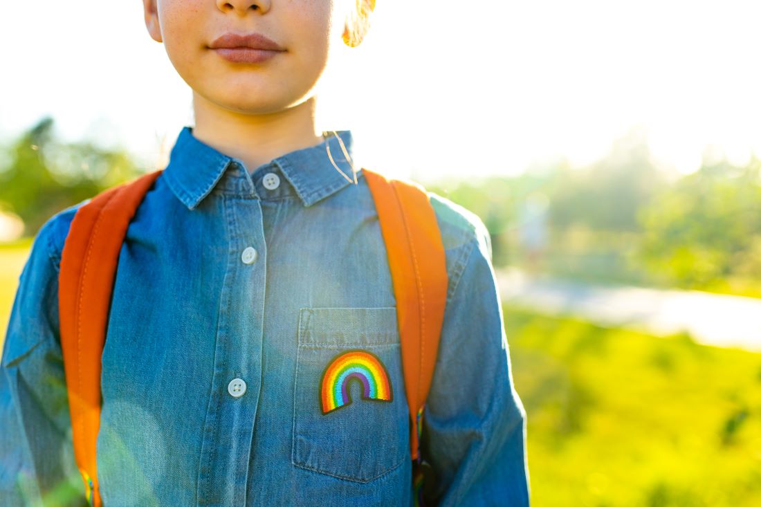child in denim t-shirt with rainbow symbol wearing orange backpack outdoors.