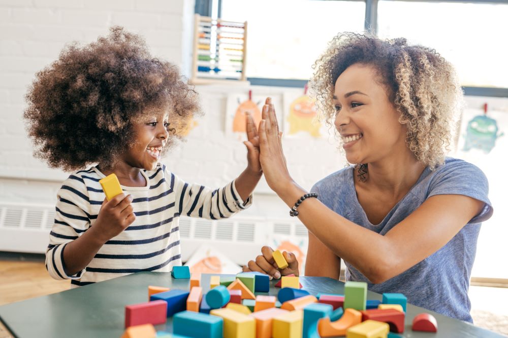 Toddler in striped shirt and caregiver in blue grey shirt play with colorful wooden blocks and give each other a high five. They both have kinky/coily hair and brown skin.