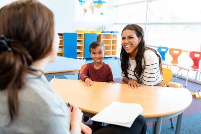 preschool age boy in a red shirt smiles as his mother laughs across the table from a teacher (the teacher's back is facing the camera and they have brown hair in a pony tail). They are seated at a table in a classroom.