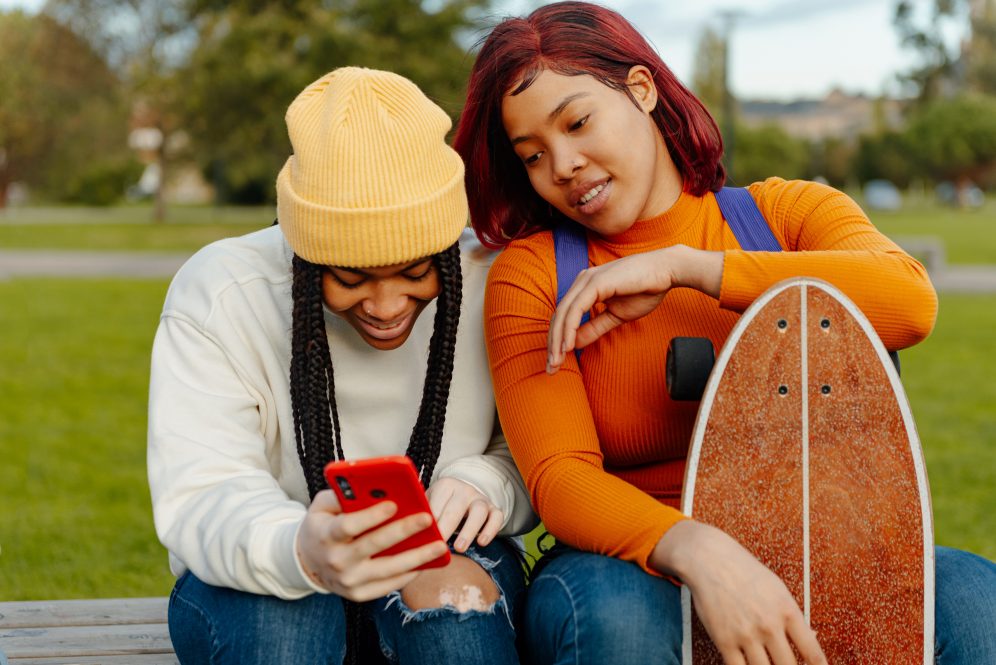 Two adolescents sit on a bench smiling at a phone