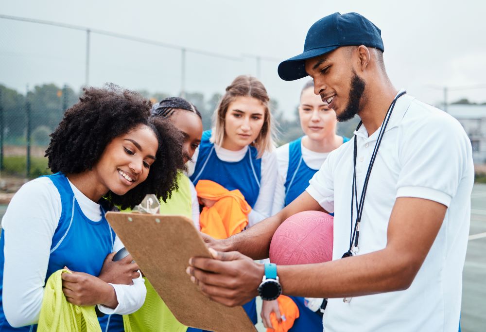 A Black male coach wearing a blue baseball cap and a whistle around his neck holds a clipboard up and shows four racially diverse girls what is on the clipboard.