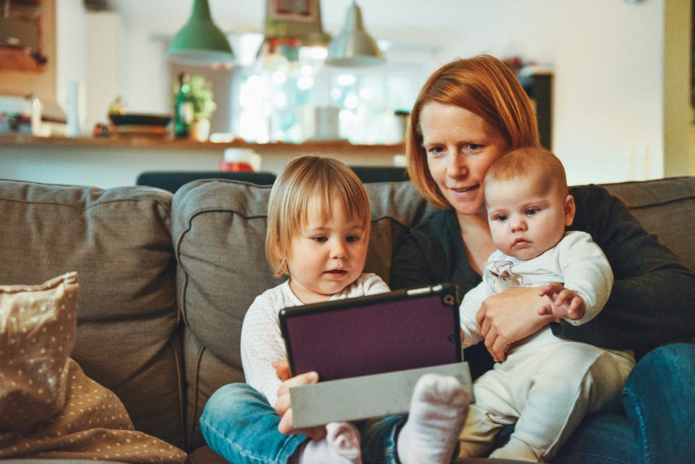 white woman with red hair sits on couch holding an infant on her lap. A toddler sits next to her holding a tablet while the woman looks at the screen