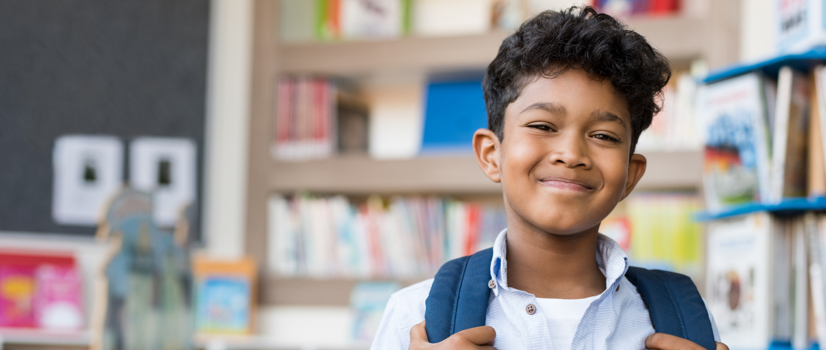 Young bow with brown skin stands in a school library with a backpack on his shoulder and smiles at the camera