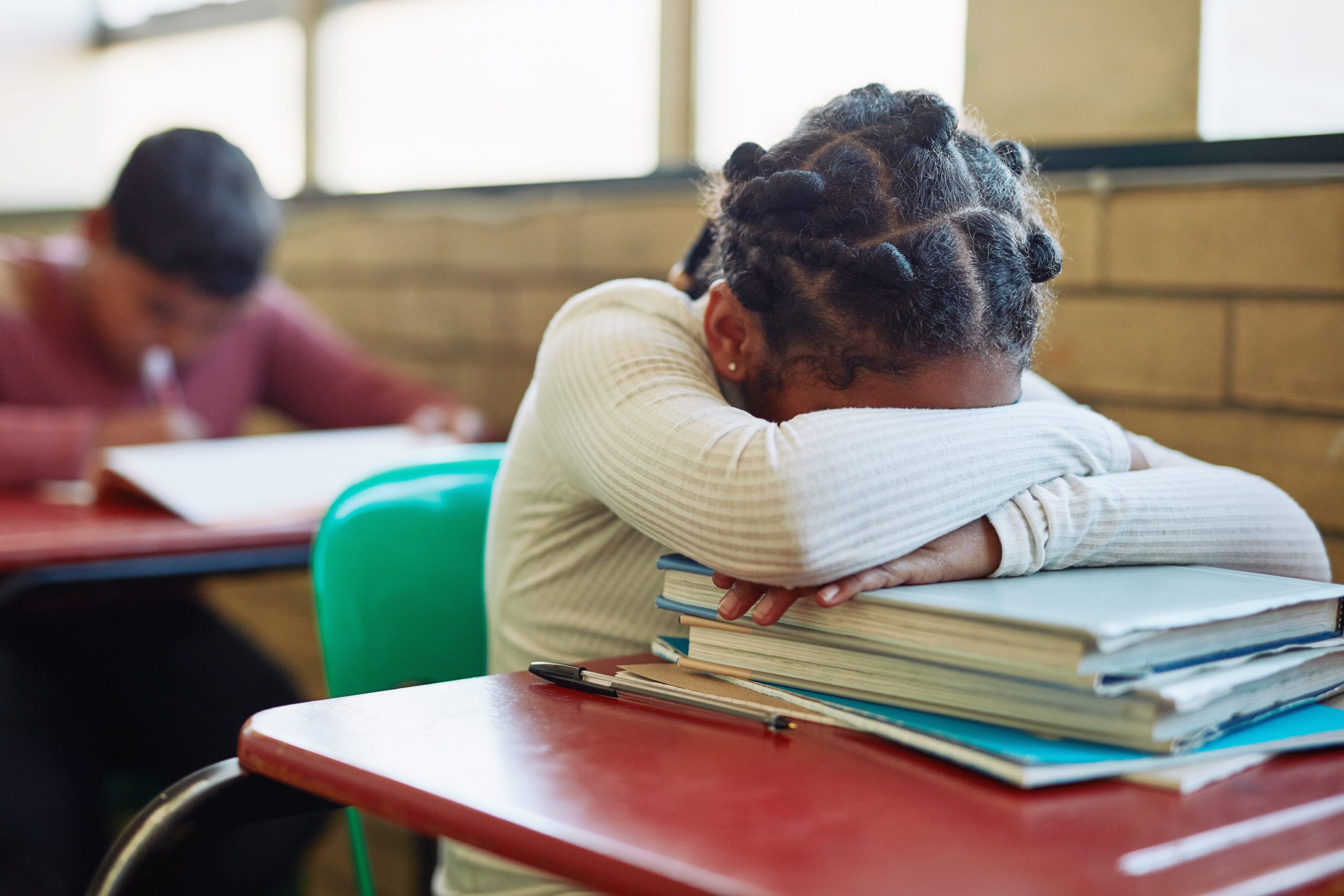 Black girl wearing a cream shirt sits at desk in classroom with her head buried in her arms.