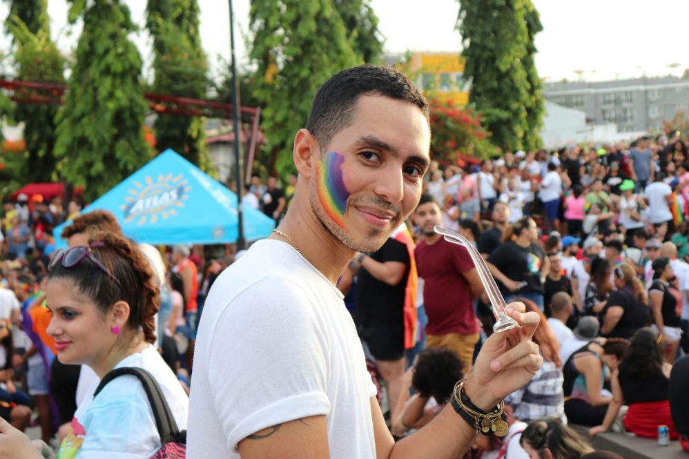 Young person smiles behind him with rainbow flag painted on his face. They hold eyeglasses in their hand. Photo by Betzy Arosemena for Unspalsh