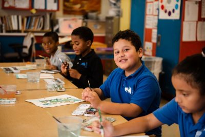 Three students sit at a desk painting. A boy on the end faces the camera and smiles