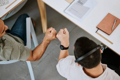 Two colleagues sit at a desk and give each other a fist bump, symbolizing teamwork and support. Office supplies, including a notebook, pen, and laptop, are visible on the desk from an overhead perspective.