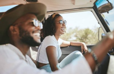 Two people are smiling and laughing while riding in a car on a sunny day. The woman in the passenger seat is wearing sunglasses, a white shirt, and a headband, and appears to be enjoying the moment. The man driving is also wearing sunglasses and a wide-brimmed hat. Both seem relaxed and happy, with bright sunlight streaming through the windows and a scenic, natural landscape outside.