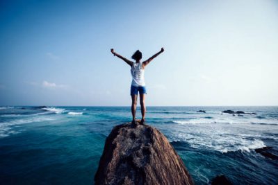 A person stands confidently atop a large rock on the edge of the ocean, arms raised in a victorious gesture. They are dressed in a white tank top and blue shorts, facing the horizon. Waves crash dramatically against the rock, and the sky above is mostly clear with a few scattered clouds, evoking a sense of adventure and triumph.