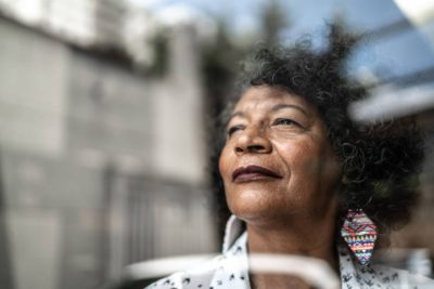 A person with curly hair wearing a white shirt patterned with small black shapes and a vibrant, multicolored triangular earring, standing outdoors against a blurred backdrop of trees and buildings.