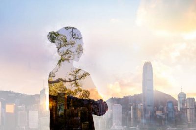 A double exposure image blending the silhouette of a person with their arms crossed and a natural forest scene of trees and foliage. Behind them is a city skyline dominated by tall buildings and a distinctive skyscraper, all set beneath a partly cloudy sky tinged with golden sunlight, suggesting dawn or dusk. The juxtaposition of urban and natural elements gives the image a reflective, harmonious feel.