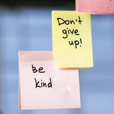 Yellow and pink sticky notes on glass with handwritten messages: 'Don't give up!' and 'Be kind'.
