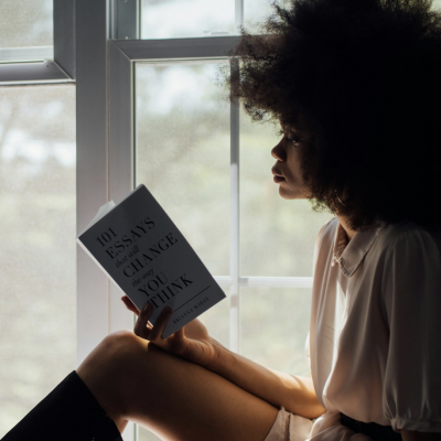 Person with curly hair reading '101 Essays That Will Change The Way You Think' by a window with natural light.