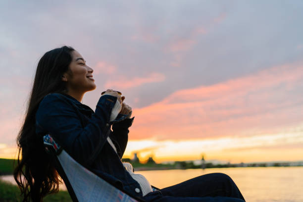 A young woman is enjoying sitting near a lake and drinking a hot drink in nature during sunset.