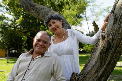 An older couple is outdoors. The man is sitting, wearing a light-colored shirt with dark buttons, while the woman stands behind him, resting her hand on a tree trunk. She is dressed in a white dress and has short gray hair. The background features lush green trees and grass, with sunlight filtering through the foliage.
