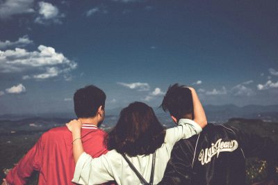 Three people stand with their backs to the camera, facing a scenic mountain landscape under a cloudy sky. The middle person has their arms around the shoulders of the other two, suggesting a close bond.
