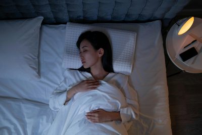 A person lies in bed on a white quilted pillow, covered by a white blanket. Their hands rest on the blanket. A bedside table on the right holds a lit lamp, a phone, and a journal with a pen.