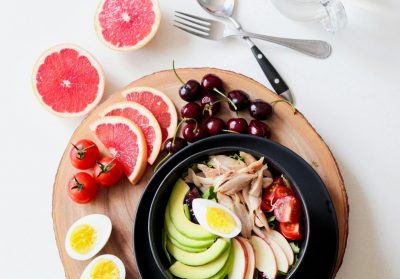 A wooden platter displays a colorful assortment of fresh foods and a salad. The salad contains avocado slices, hard-boiled egg halves, shredded chicken, cherry tomatoes, and apple slices. A fork, spoon, and an empty glass jar are placed above the platter.