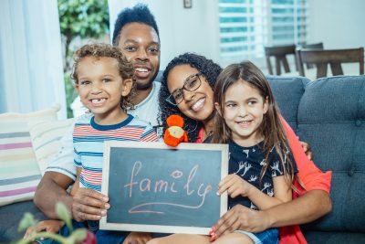 A family of four is sitting on a couch in a living room, holding a small chalkboard with the word 'family' written on it in cursive. The background includes a window with blinds and some furniture.