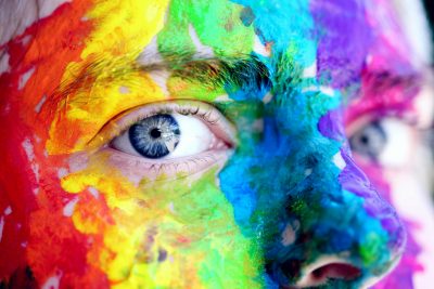 A close-up of a person's face covered in bright, multicolored paint. The paint is applied in a random, artistic pattern with vivid shades of red, orange, yellow, green, blue, and purple. The person's blue eye is clearly visible and sharply in focus, standing out against the colorful background.