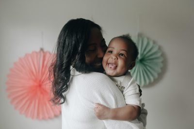 A person with long hair holding a baby, both are smiling.