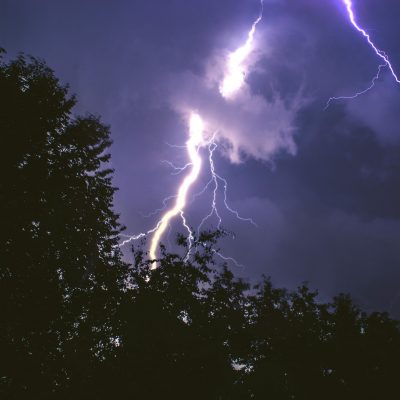 A nighttime scene with a dark sky lit up by several bright lightning bolts. Tree silhouettes are visible in the foreground, and the lightning appears to strike through and around them. The sky features a mix of dark clouds and glowing areas from the lightning.
