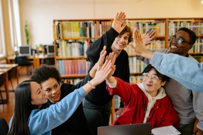 A group of six people in a library are gathered around a table, raising their hands together in a high-five gesture. The background includes bookshelves filled with books and several computer stations.